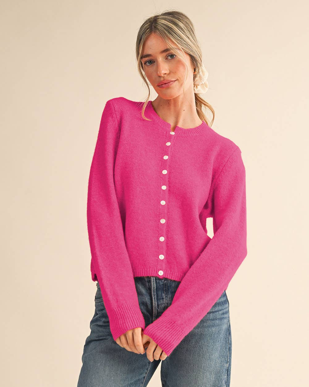 A woman modeling a bright pink knit cardigan sweater with white buttons down the front, paired with blue jeans, against a solid beige background.