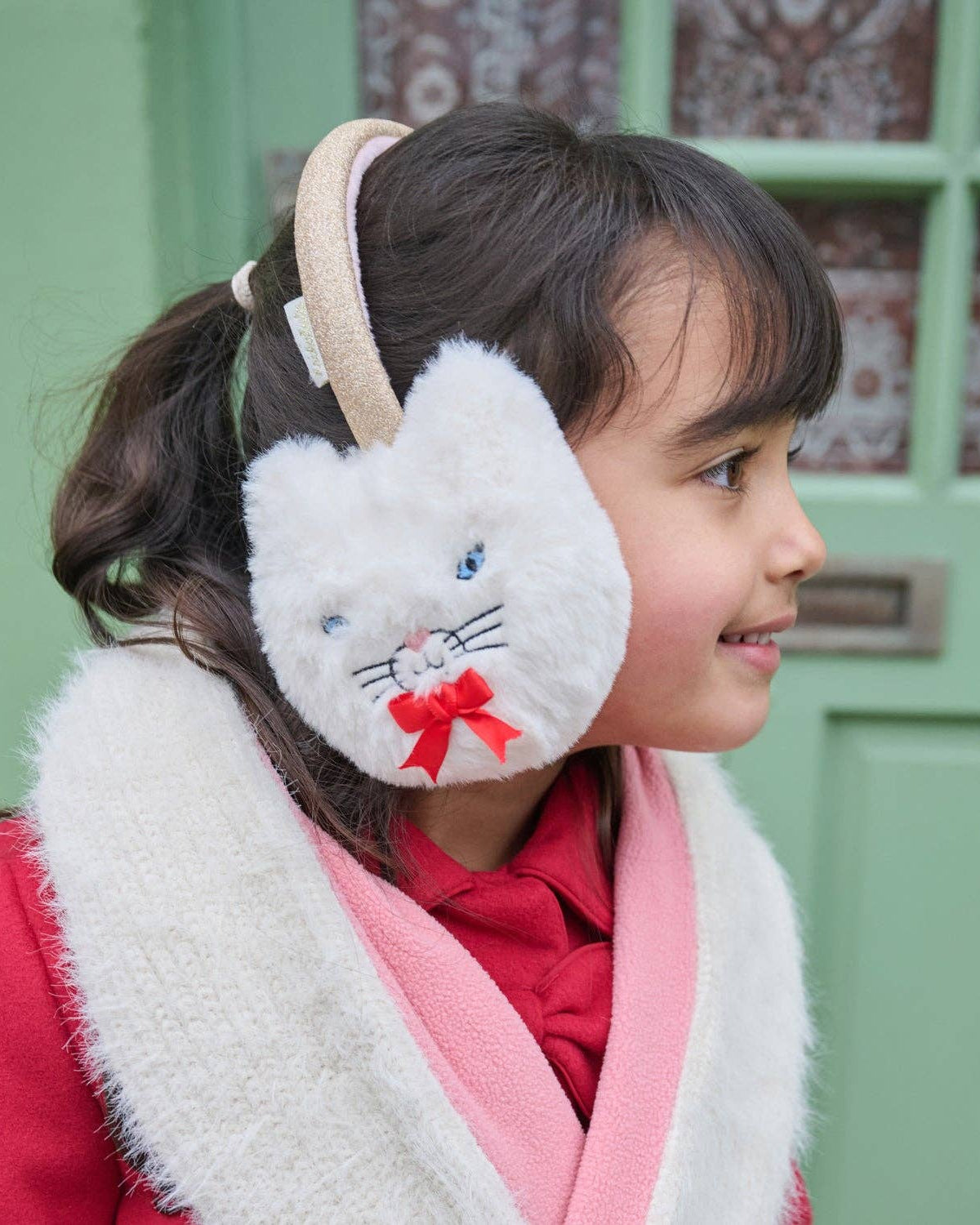 A young girl wearing white cat-ear earmuffs with a red bow and a pink-and-white knit scarf, shown in profile against a green door.