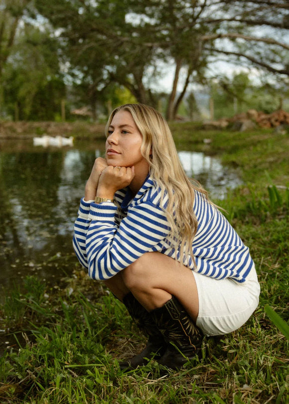Woman in a striped shirt and white skirt squatting by a pond with trees in the background