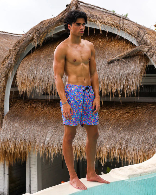 Man wearing colorful swim shorts standing by a pool with thatched-roof structures in the background