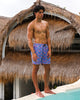 Man wearing colorful swim shorts standing by a pool with thatched-roof structures in the background