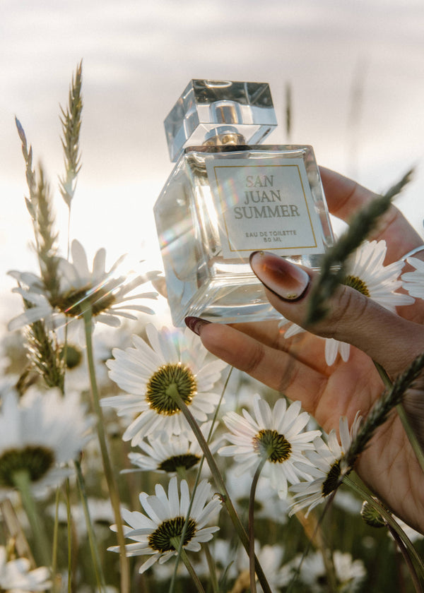 Hand holding a perfume bottle labeled 'San Juan Summer' amidst white flowers