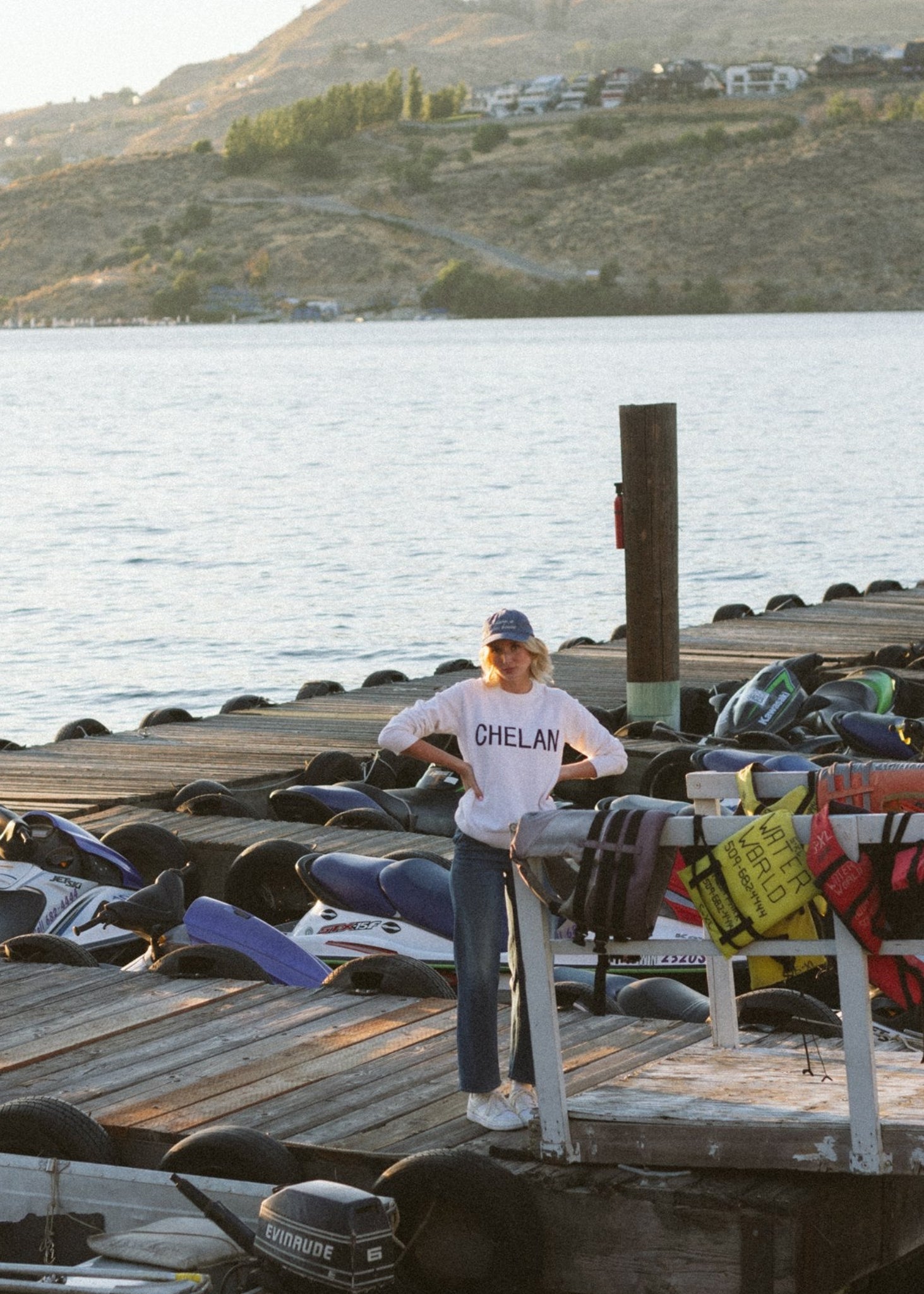 A person wearing a white knit sweater standing on a dock with boats and lake in the background.