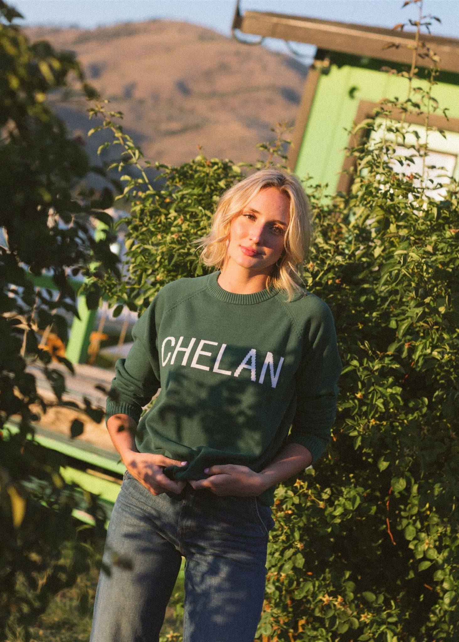 A person wearing a green crew neck sweater with 'CHELAN' written across the front in white letters, standing near greenery with hills in the background.
